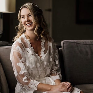 May include: A woman wearing a white lace robe with floral embroidery sits on a gray couch. She is smiling and looking to the side. She is wearing a tiara and earrings.