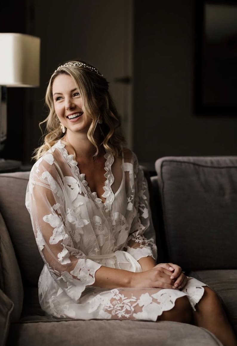May include: A woman wearing a white lace robe with floral embroidery sits on a gray couch. She is smiling and looking to the side. She is wearing a tiara and earrings.