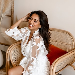 May include: A woman wearing a white lace robe with floral embroidery sits in a wicker chair with a red cushion. She is smiling and looking at the camera.