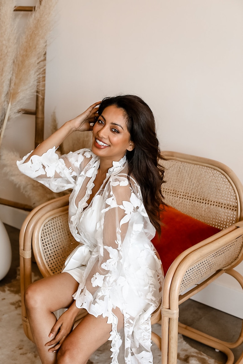 May include: A woman wearing a white lace robe with floral embroidery sits in a wicker chair with a red cushion. She is smiling and looking at the camera.