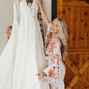 Puede incluir: Una mujer con una bata blanca de flores sostiene un vestido de novia blanco. Ella sonr&iacute;e y mira el vestido. El vestido est&aacute; colgado en una percha frente a una puerta de madera.