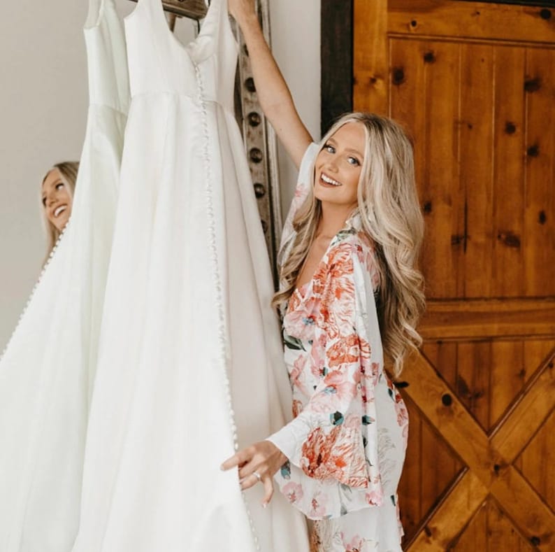 Puede incluir: Una mujer con una bata blanca de flores sostiene un vestido de novia blanco. Ella sonr&iacute;e y mira el vestido. El vestido est&aacute; colgado en una percha frente a una puerta de madera.
