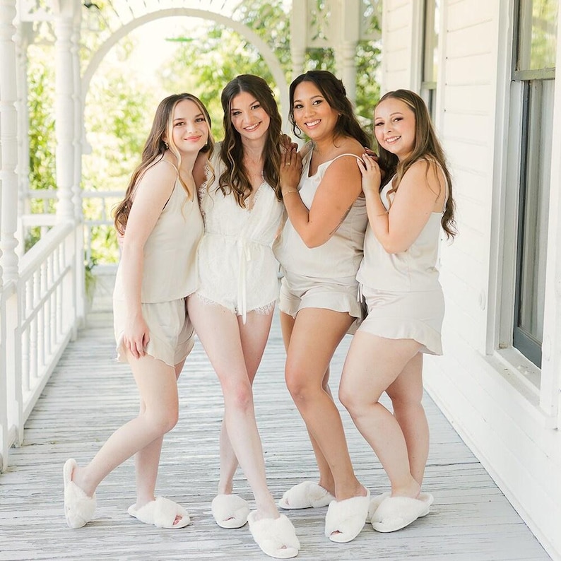 May include: Four people wearing matching beige sleepwear, including shorts, tank tops, and a romper. They are also wearing white fluffy slippers. The group is standing on a white porch with a white railing and a green background.