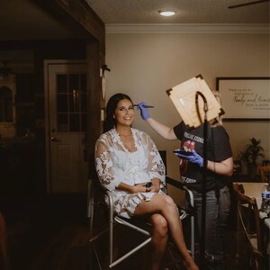 May include: A woman in a white lace robe sits in a chair while another woman applies makeup to her face. The woman applying makeup is wearing a black t-shirt with the words "Rolling Stone" on it.