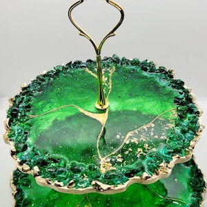 May include: Two-tiered serving tray with a gold handle and trim. The trays are a vibrant green with gold accents and a textured, gem-like border. A decorative piece for displaying desserts or small items.
