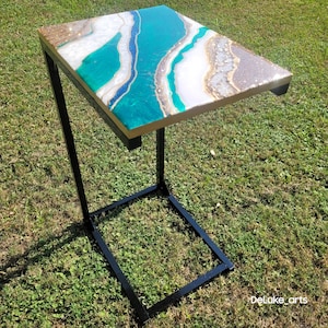 May include: A side table with a rectangular top featuring a teal, white, and gold resin design. The table has a black metal frame and a single leg. The table is approximately 24 inches long and 12 inches wide.