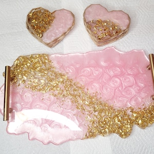 May include: Pink resin tray with gold handles and two heart-shaped coasters. The tray and coasters are pink with gold leaf accents. The tray has a wavy edge and a swirled pattern.
