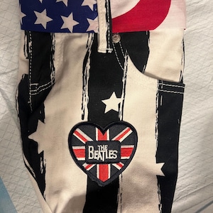 May include: A fabric bag with a design resembling denim jeans. The bag features a black and white striped pattern, a pocket, and a fold-over top with a blue and white star pattern and red stripes. A heart-shaped patch with "The Beatles" text is sewn on.