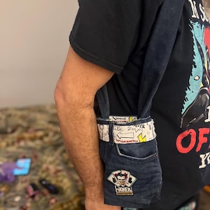 Puede incluir: Un bolso cruzado de mezclilla hecho de jeans reciclados. El bolso presenta un bolsillo con un parche de "Joker" y una correa blanca tipo cinturón con gráficos de estilo cómic y el texto "Captain America".