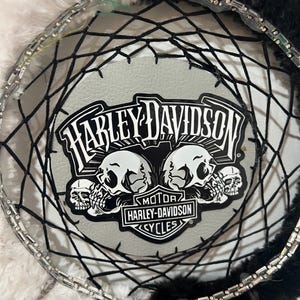 May include: A Harley-Davidson dreamcatcher with a black and white color scheme. The center features the Harley-Davidson logo with skulls. The dreamcatcher has a beaded rim and black and white feathers.