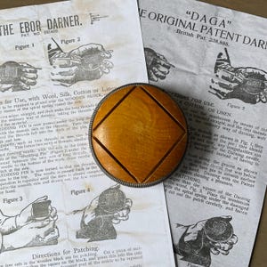 May include: A wooden darning mushroom with a metal rim rests on old instruction sheets. The mushroom has a square shape with a diamond design. The papers display illustrations and text about darning techniques, with the text "THE EBOR DARNER" and "DAGA".