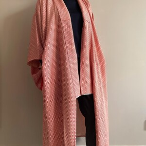 May include: A long, red and white patterned kimono with a black turtleneck underneath. The kimono has a wide, open collar and long sleeves.