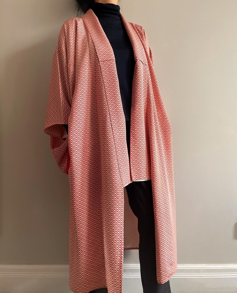 May include: A long, red and white patterned kimono with a black turtleneck underneath. The kimono has a wide, open collar and long sleeves.