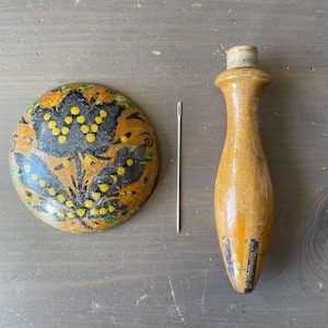 Puede incluir: Un estuche de agujas de madera y un cojín decorativo con un diseño floral. El cojín es redondo con un patrón floral negro, amarillo y naranja. El estuche de agujas es de color marrón claro con un tapón de corcho.
