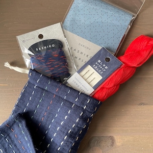 May include: A navy blue drawstring bag filled with Sashiko embroidery supplies. The bag is decorated with white, red, and green stitching. Included are spools of thread, needles, and fabric samples. A bundle of red thread is also visible.