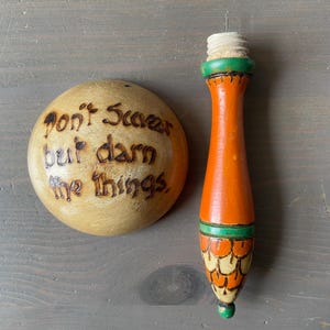 Könnte beinhalten: Ein hölzerner Flaschenverschluss und ein runder Holzgegenstand. Der Flaschenverschluss ist orange mit grünen und beigen Akzenten. Der runde Gegenstand trägt den Text "Don't Scare but darn the things" in das Holz gebrannt.