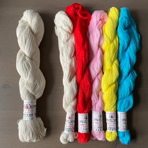 May include: Six skeins of embroidery floss in various colours, including white, red, pink, yellow, and blue. Each skein has a label with Japanese text and the word "ダルマ" (Daruma).