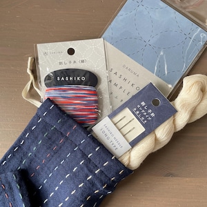 Könnte beinhalten: Ein marineblauer Stoffbeutel mit weißen Nähten, der Sashiko-Garn, Nadeln und Musterbeispiele enthält. Das Garn ist rot, weiß und blau. Die Verpackung enthält japanischen Text und den Markennamen "DARUMA."