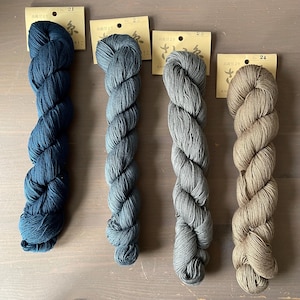 May include: Four skeins of yarn in varying shades of blue, gray, and tan. Each skein is labeled with a number and Japanese text. The yarn is tightly twisted, showcasing its texture and color variations. The skeins are arranged on a wooden surface.