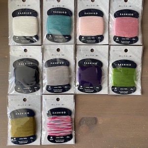 May include: A selection of Sashiko thread spools in a range of colours: white, teal, pink, rose, black, grey, purple, and green. Each spool is labelled with the brand name and product details. The thread is 100% cotton, 40m.