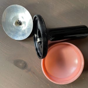 May include: A collection of vintage camera components, featuring a silver reflector, a black cylindrical body, and a pink plastic piece. The parts are displayed on a wooden surface, highlighting their distinct forms and textures.