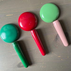 May include: Three brightly coloured, round-topped items with cylindrical handles. One is red, one is green, and one is pink. The handles are attached to the round tops. They are arranged on a grey wooden surface.