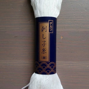 May include: A spool of white embroidery thread with a navy blue label featuring Japanese text and a gold wave pattern. The thread is tightly wound and ready for use in various needlework projects.