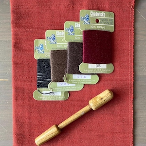 May include: Four cards of Chadwicks wool and nylon mending yarn in black, brown, dark brown, and burgundy, each labelled 10m 10.9yd. A wooden darning tool with a cylindrical handle and a thicker head rests below the yarn cards on a red fabric.