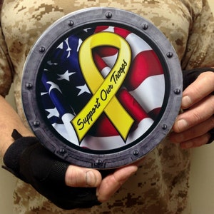 May include: A round metal sign with a yellow ribbon and an American flag. The ribbon has the words "Support Our Troops" written on it.