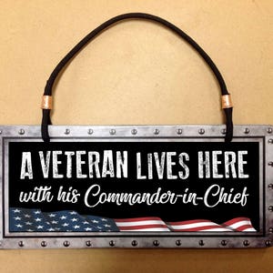 May include: A metal sign with a black background and white text that reads "A Veteran Lives Here with his Commander-in-Chief". The sign has a distressed look and features a red, white, and blue American flag.