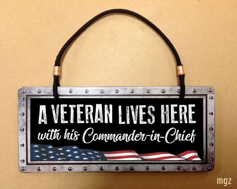May include: A metal sign with a black background and white text that reads "A Veteran Lives Here with his Commander-in-Chief". The sign has a distressed look and features a red, white, and blue American flag.