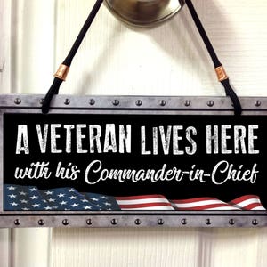 May include: A metal sign with a distressed look, featuring a black background with white text that reads "A Veteran Lives Here with his Commander-in-Chief". The sign has a red, white, and blue American flag design at the bottom.
