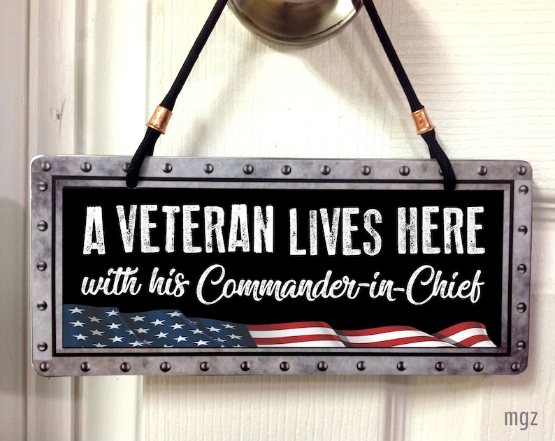 May include: A metal sign with a distressed look, featuring a black background with white text that reads "A Veteran Lives Here with his Commander-in-Chief". The sign has a red, white, and blue American flag design at the bottom.
