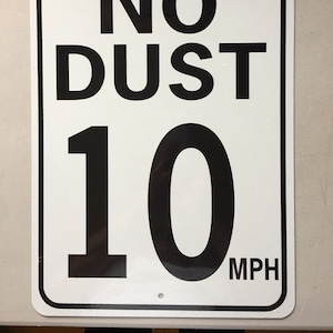 Op de afbeelding: Wit metalen bord met zwarte tekst die "PLEASE... NO DUST 10 MPH" luidt.