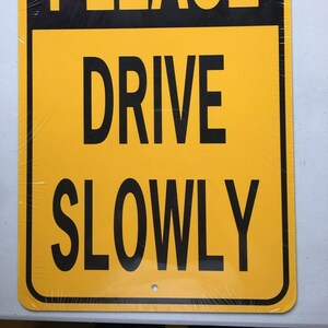 Op de afbeelding: Een geel en zwart bord met de tekst "PLEASE DRIVE SLOWLY" in zwarte letters.