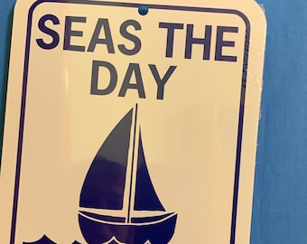 May include: White metal sign with blue text and a sailboat graphic. The sign reads "Seas the Day".