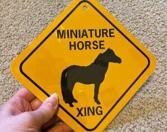 Op de afbeelding: Een geel waarschuwingsbord met zwarte tekst en een silhouet van een miniatuurpaard. Het bord luidt "Miniature Horse Xing".