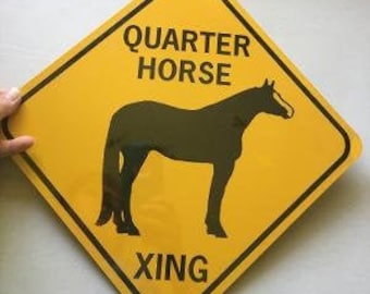 Op de afbeelding: Geel ruitvormig verkeersbord met zwarte tekst en een zwart silhouet van een paard. Het bord luidt "Quarter Horse Xing".