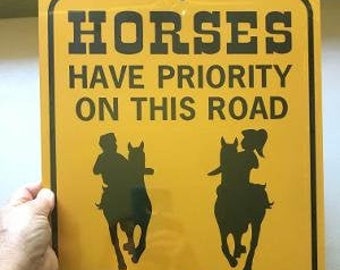 Op de afbeelding: Geel verkeersbord met zwarte tekst die "Horses Have Priority On This Road Slow Down" luidt. Twee zwarte silhouetten van paarden en ruiters zijn op het bord afgebeeld.