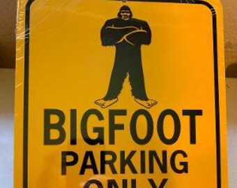 May include: Yellow Bigfoot parking sign with black text and a black Bigfoot graphic. The sign reads "BIGFOOT PARKING ONLY" with arrows and "ALL OTHERS WILL BE STOMPED". The sign is rectangular with rounded corners.