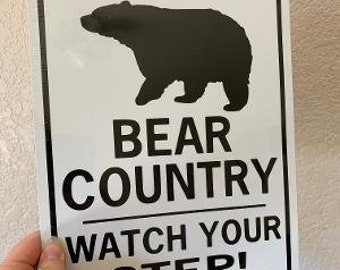 Op de afbeelding: Een rechthoekig wit bord met een zwarte rand. Het bord toont een zwart silhouet van een beer bovenaan, met de woorden "BEAR COUNTRY" en "WATCH YOUR STEP!" in vette zwarte letters.