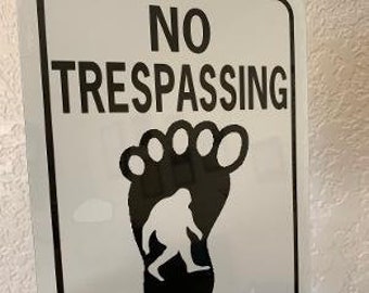 May include: A white metal sign with black text that reads "NO TRESPASSING". A black footprint with a white silhouette of a Bigfoot inside is below the text.