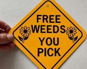 Op de afbeelding: Een geel ruitvormig bord met zwarte tekst die "FREE WEEDS YOU PICK" luidt. Twee gestileerde zwart-witte bloemenillustraties bevinden zich aan weerszijden van de tekst.
