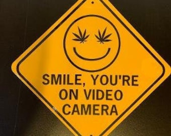 Op de afbeelding: Geel ruitvormig bord met een zwarte rand. Het heeft een lachend gezicht met cannabisbladeren als ogen. De tekst "SMILE, YOU'RE ON VIDEO CAMERA" is in het zwart gedrukt.