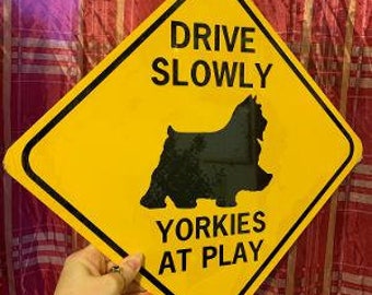 May include: Yellow diamond-shaped sign with black text and a black silhouette of a Yorkshire Terrier. The sign reads "Drive Slowly Yorkies At Play".