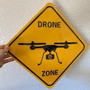 Puede incluir: Señal amarilla en forma de diamante con texto negro y una silueta negra de un dron. La señal dice "Drone Zone".