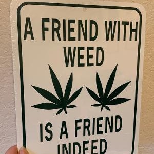 May include: A rectangular sign with rounded corners features the text "A FRIEND WITH WEED IS A FRIEND INDEED" in black. Two green cannabis leaves are centered above the words "IS A FRIEND". The sign has a white background and a green border.