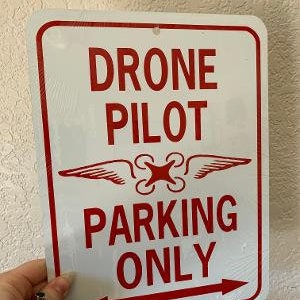 Puede incluir: Un letrero de metal blanco con texto rojo que dice "Drone Pilot Parking Only". El letrero tiene una flecha roja apuntando hacia la derecha y un símbolo de dron rojo con alas.