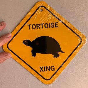 Może przedstawiać: Żółty znak w kształcie rombu z czarnym tekstem i czarną sylwetką żółwia. Znak brzmi "TORTOISE XING".