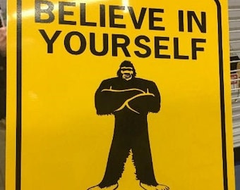 May include: Yellow metal sign with black text and a black silhouette of a Bigfoot. The sign reads "Believe in Yourself Even When Others Don't".
